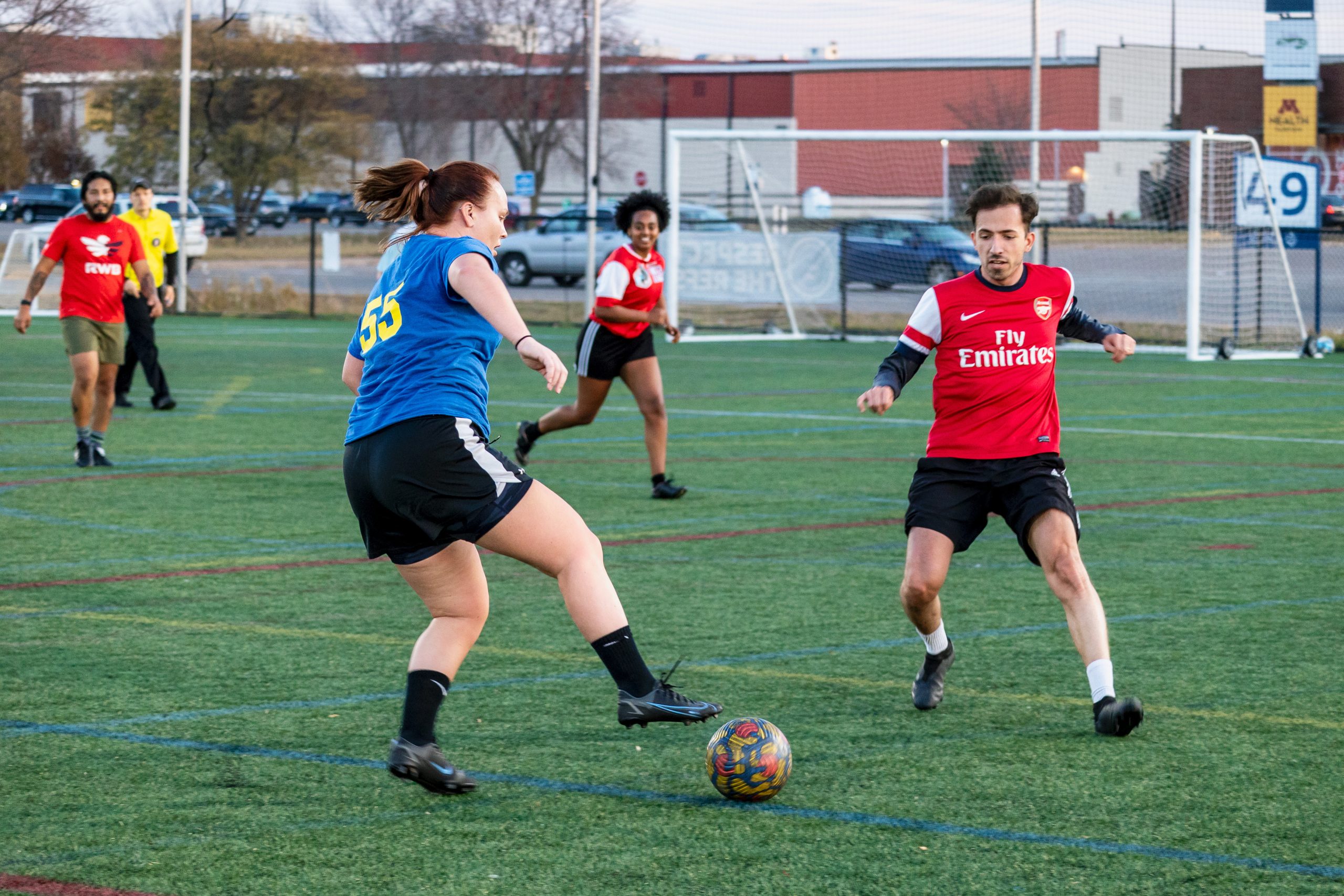 Woman in blue shirt keeping soccer ball away from man in red MNUFC Adult League shirt.