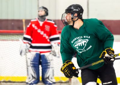 A hockey player wearing a green "Minnesota Wild Adult Hockey League" jersey, with a goalie in the background.