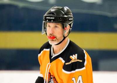 A hockey player wearing an orange and black jersey, with a helmet and red mouthguard, on an ice rink.
