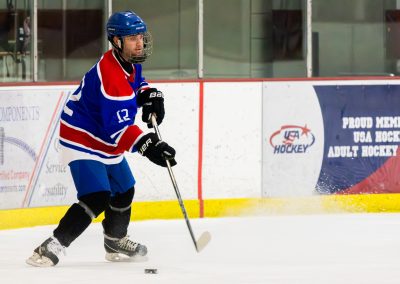 A hockey player in a blue, red, and white uniform skates on ice with a stick, with a USA Hockey banner in the background.