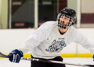A hockey player in a white jersey with a face cage is on the ice holding a stick.